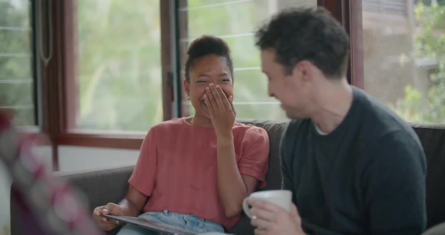 A man and a woman in a room sitting on a couch with the man drinking from a mug and the woman looking at a tablet