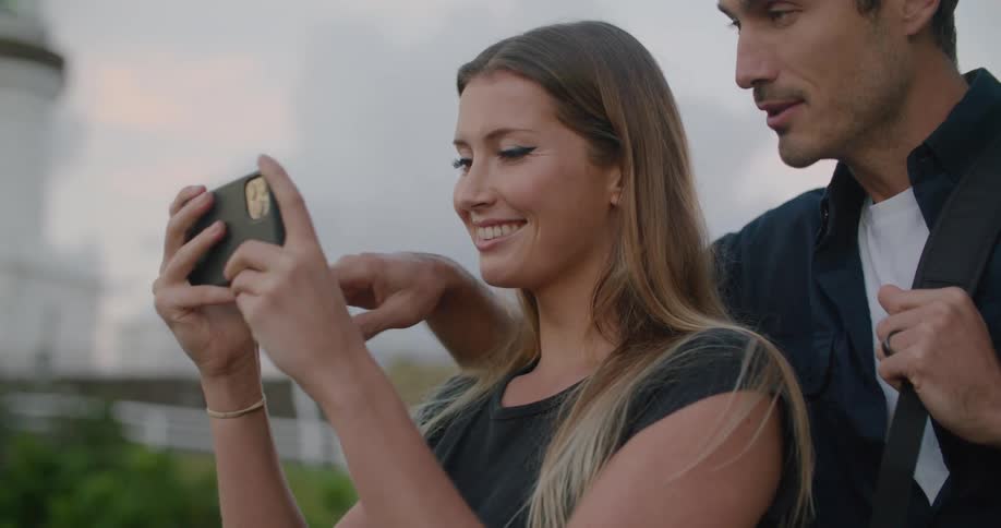 A man and a woman looking at a smart phone with a fence the sky and clouds in the background