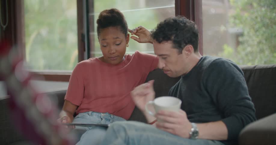 Man wearing a black sweater and jeans sits next to woman with short hair red t shirt and jeans on couch using tablet in