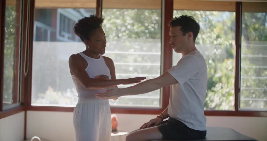 A woman stands in front of a man sitting on a medical table describing an arm turning motion to him