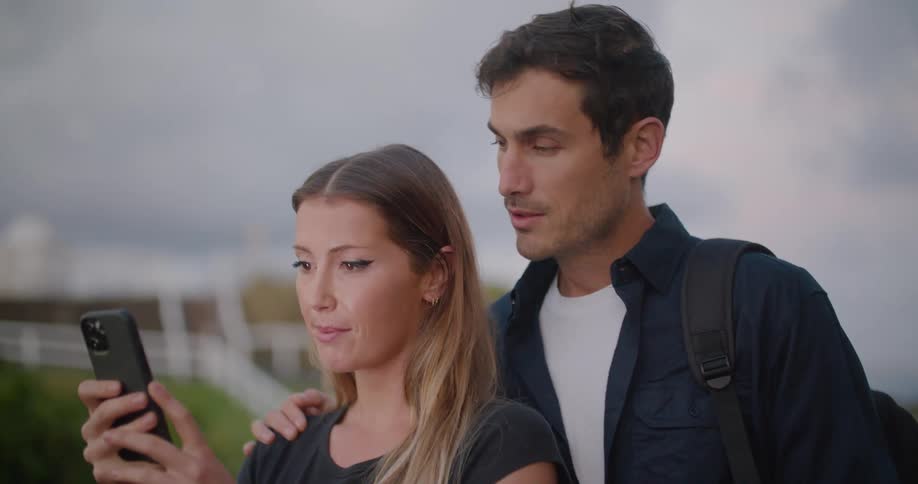A man and a woman looking at a smart phone with a fence the sky and clouds in the background