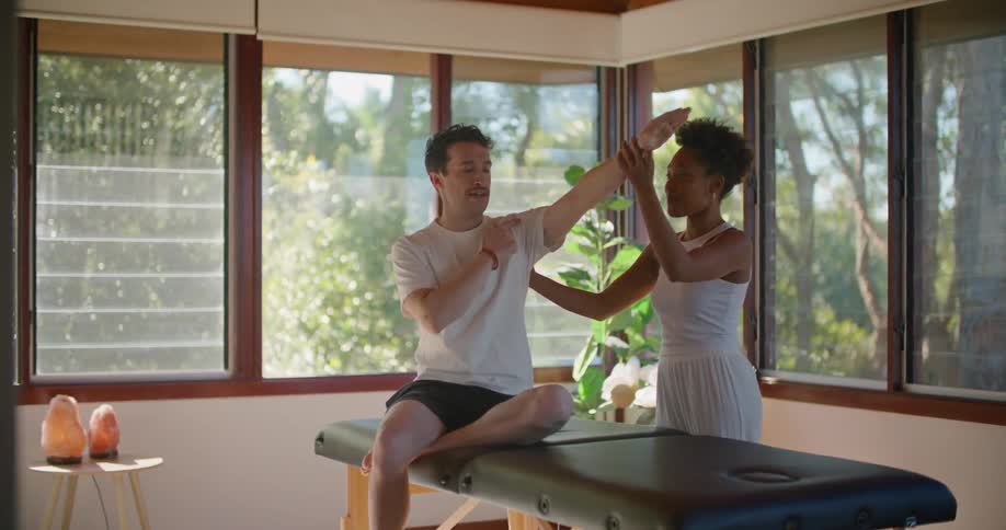 A man and a woman in a room with the man sitting on a massage table when a plant and windows in the background