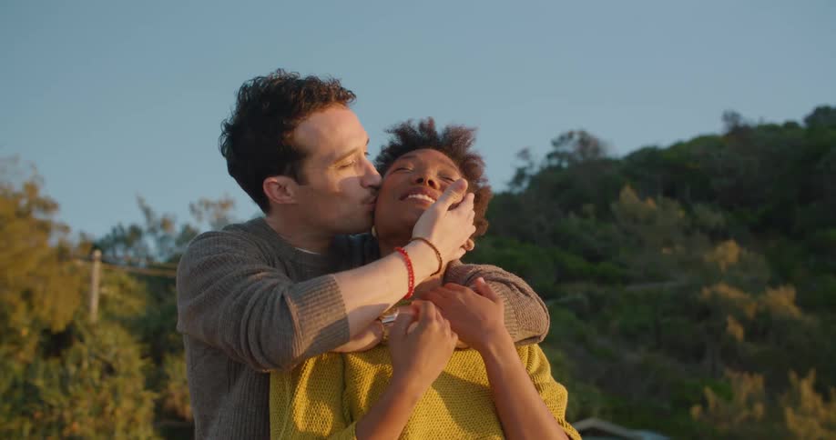 A man stands behind a woman outside and hugs and kisses her