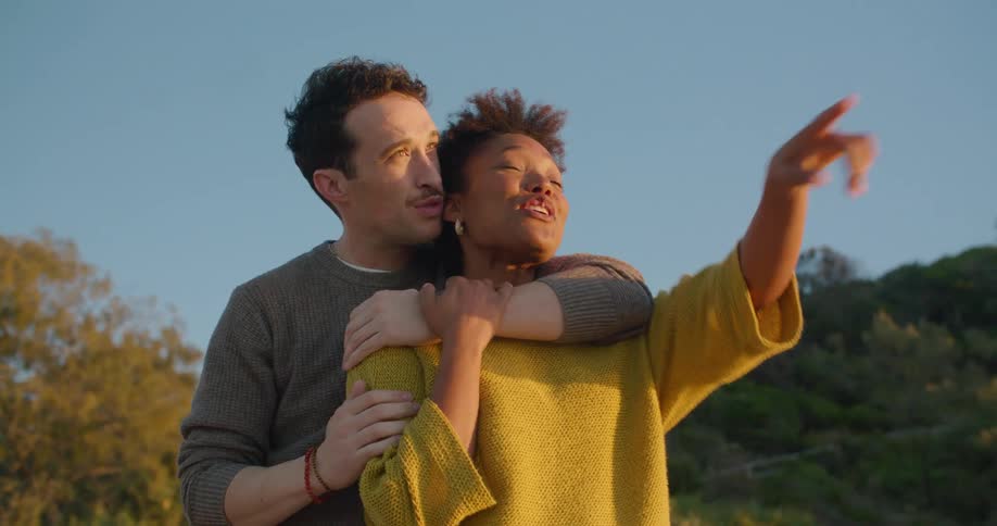 Man stand behind woman hugging as she points with hill in background