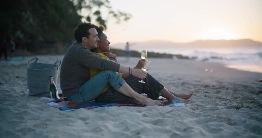 A man hugs his girlfriend to his chest while they sit on the sand holding champagne looking at the ocean