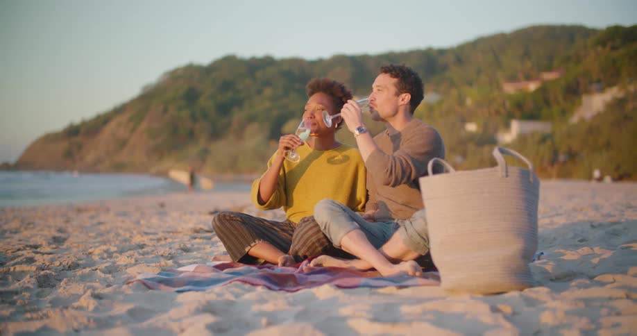 Young couple sitting on a blanket over the sand of a beach drinking while looking at the sunset