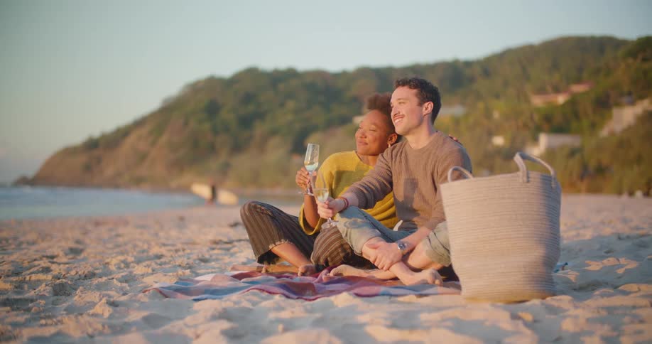 A man and woman drink wine together while sitting on the beach during the day
