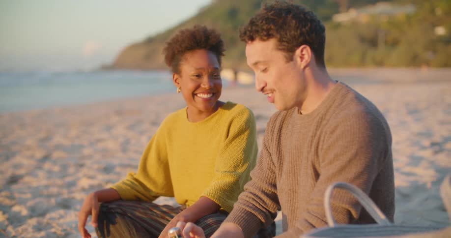 A man and woman sitting on a blanket on the beach smile and talk as he opens a bottle of champagne