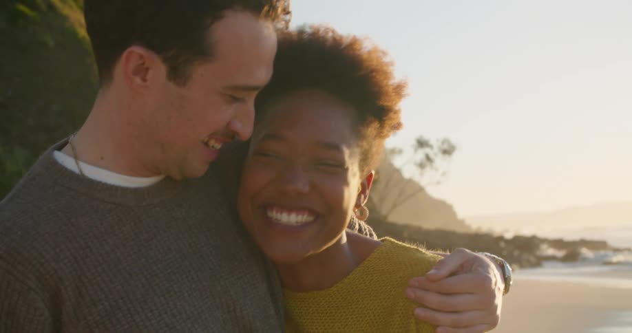 A man and woman with their arms around each other stand on the beach laughing and smiling
