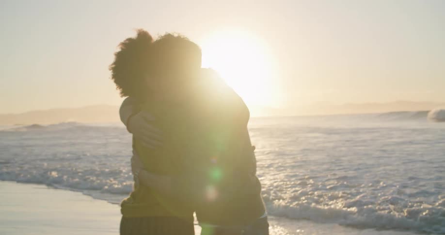 A man and woman hug on the beach near the waves then step back holding hands