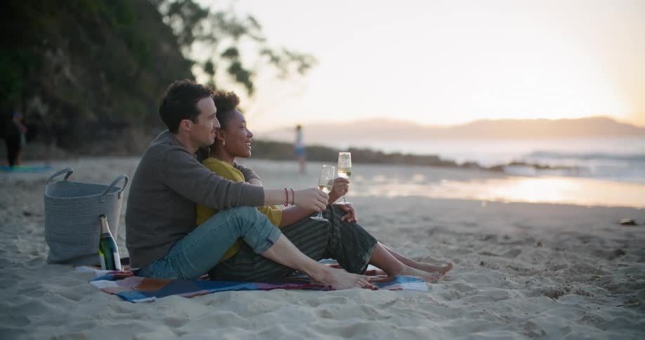 A man hugs his girlfriend from behind as they sit on the beach drinking wine and looking at the ocean