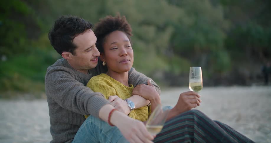 A man hugs his girlfriend from behind as they sit on the beach drinking wine
