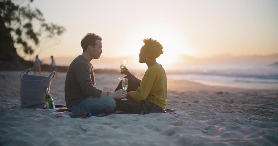 A man and woman sitting on the beach drinking wine