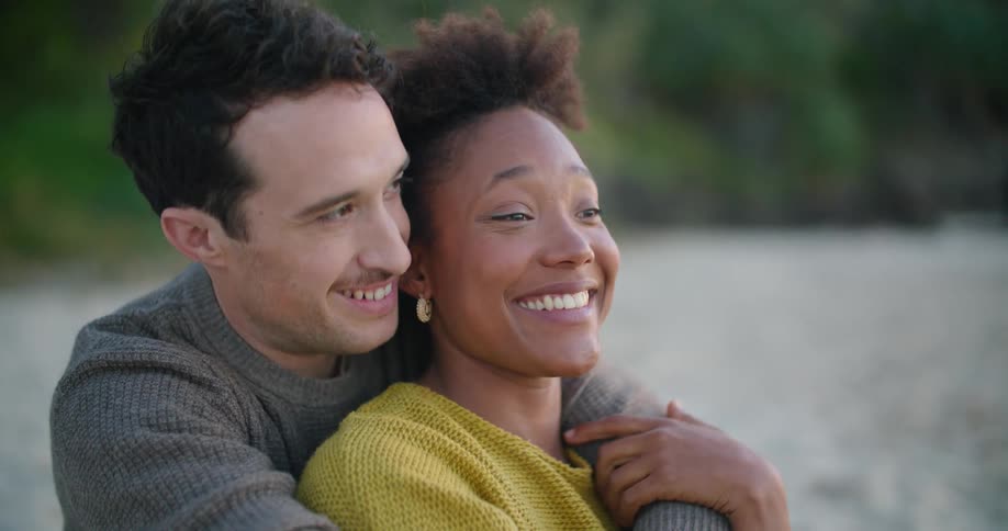 Couple sit on beach wearing sweaters with woman leaning back against man laughing and talking