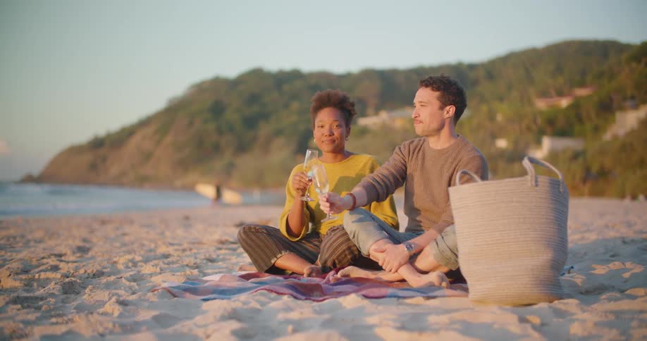 Young couple sitting on the sand of a beach toasting with wine at sunset