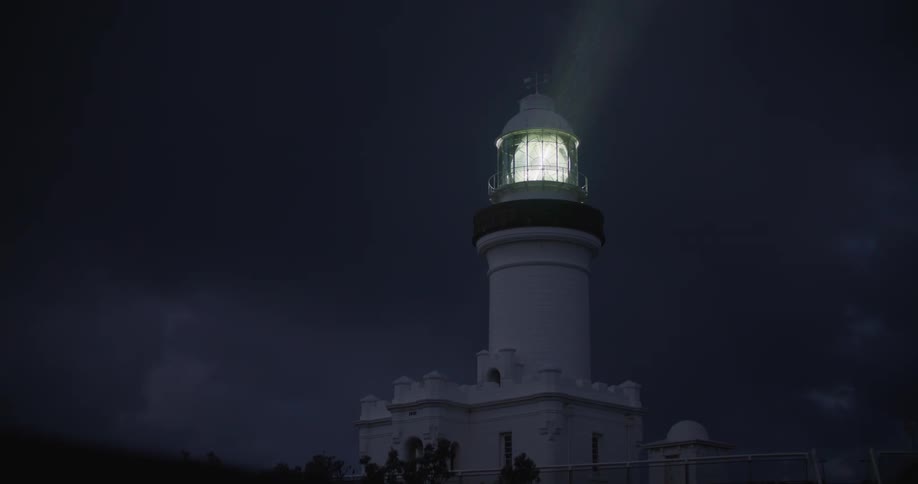 Light rotates in tower of lighthouse under cloudy sky at night