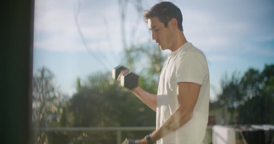 A man picks up dumbbells and does bicep curls on a deck in the sunshine