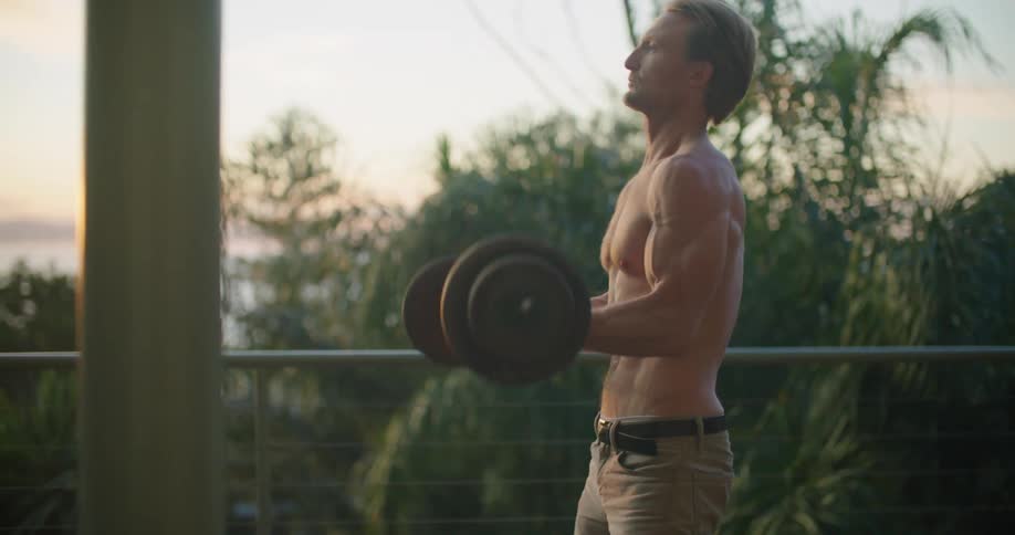 Shirtless muscular man lifting weights outside the house at sunset