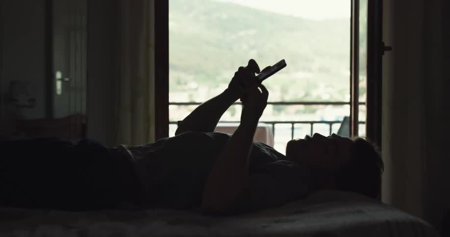 Man lying down on bed uses smartphone and is silhouetted against the balcony
