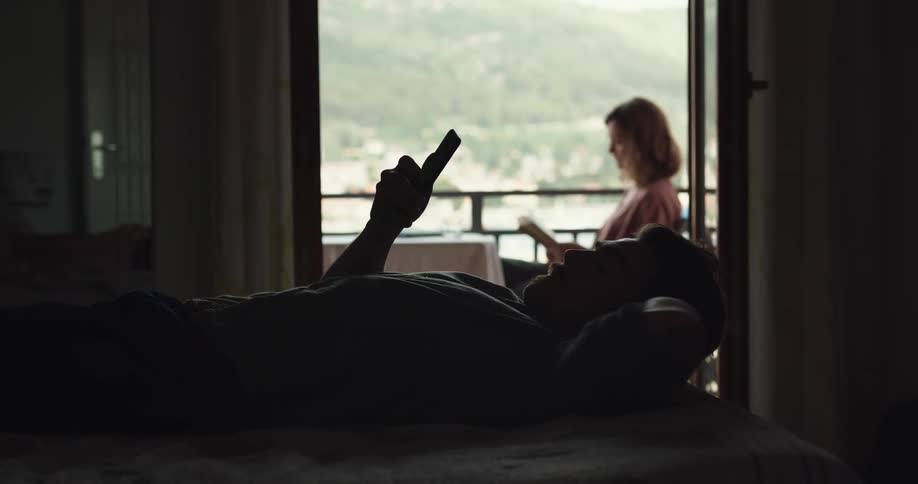 A man leisurely views his smartphone while lying on a bed in a dark room as a woman reads a book on a nearby balcony