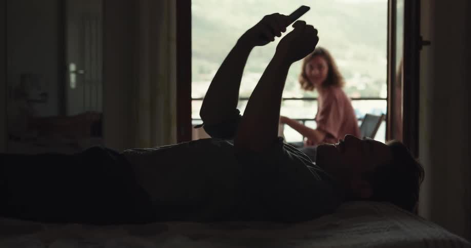 Man lying down on couch uses smartphone and woman comes up to man to cuddle and kiss