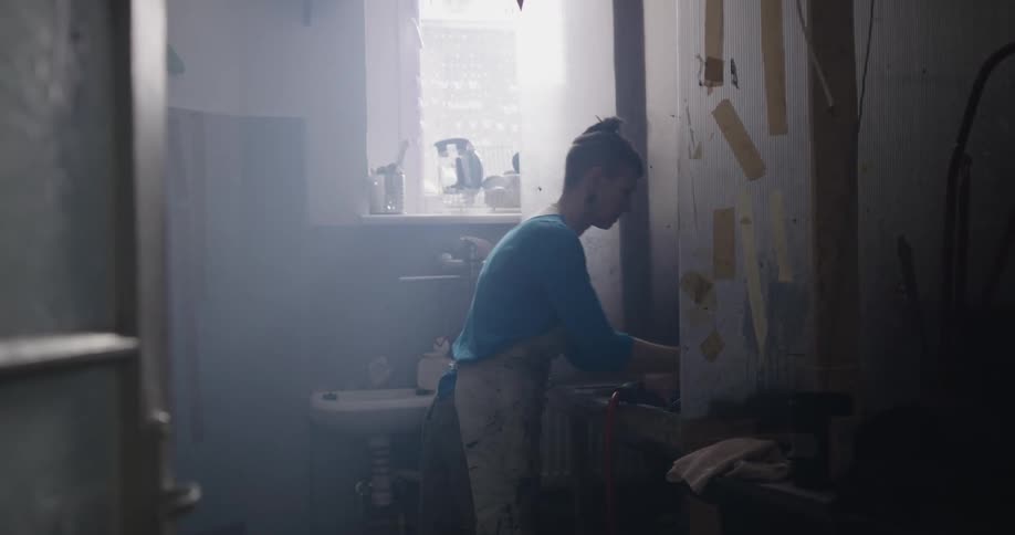 Woman walks across dimly lit room to open paint can on work stand