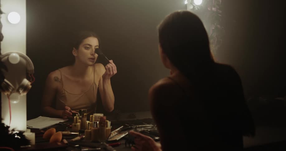 A woman sits in front of a mirror looking at papers then stretching and using a makeup brush on her face