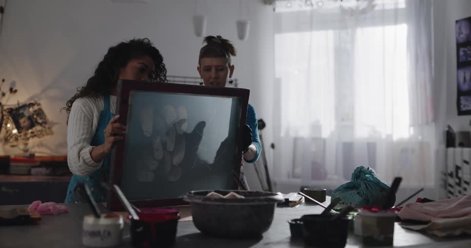 Two women stand in an art studio looking at a framed piece of art