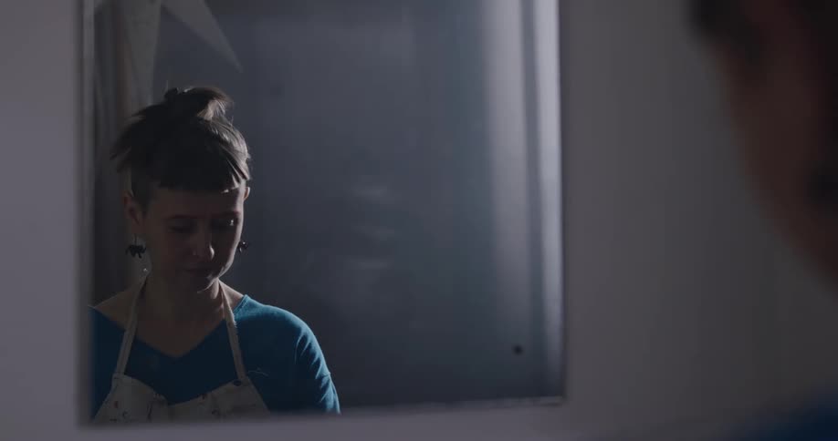 A young woman in an apron looks down as she stands in front of a mirror