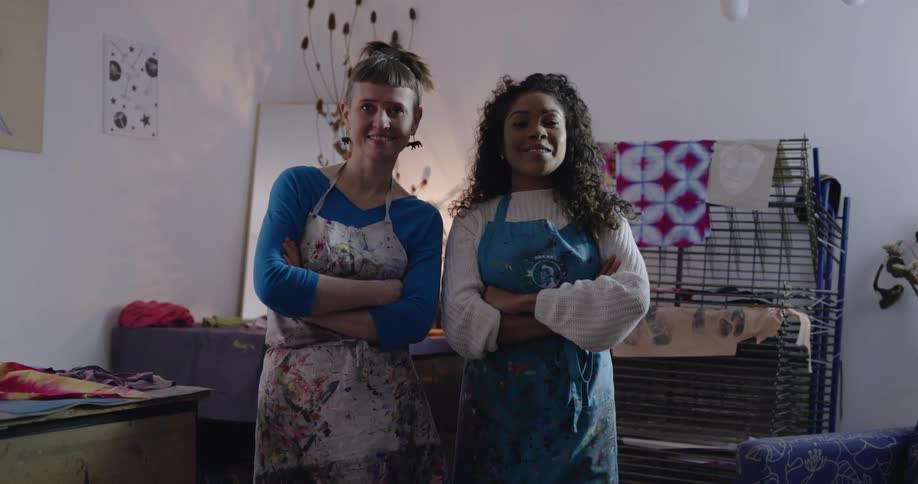 Two women wearing aprons stand in front of some fabric on a rack smiling with their arms folded