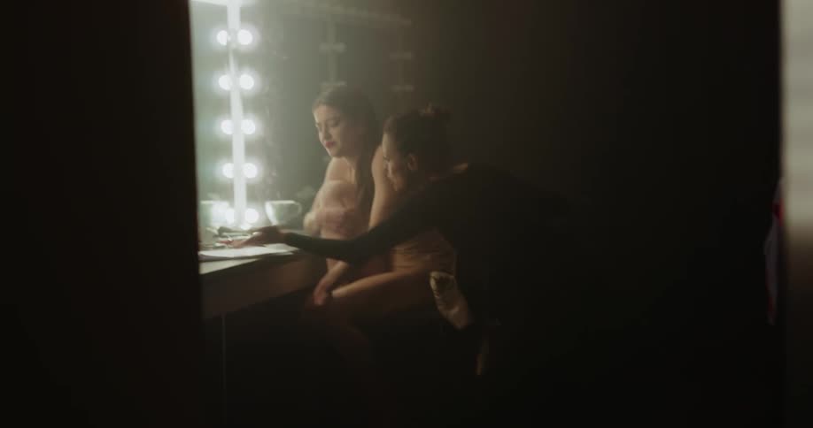 Older woman wearing leotard walks into dressing room and yells at dancer sitting in front of mirror over bundle of papers