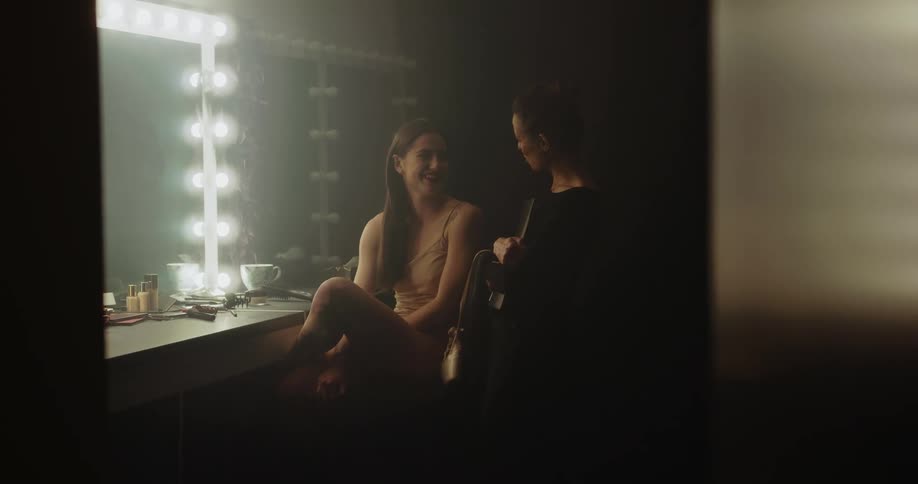 Two women in a dressing room with one woman holding a clipboard with a wall and a mirror in the background