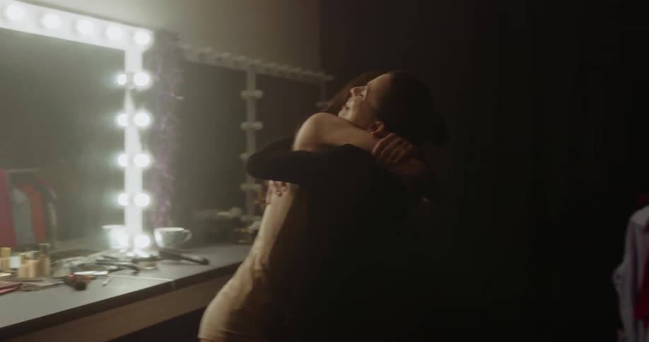 Two women in a dressing room hugging with a mirror and a wall in the background