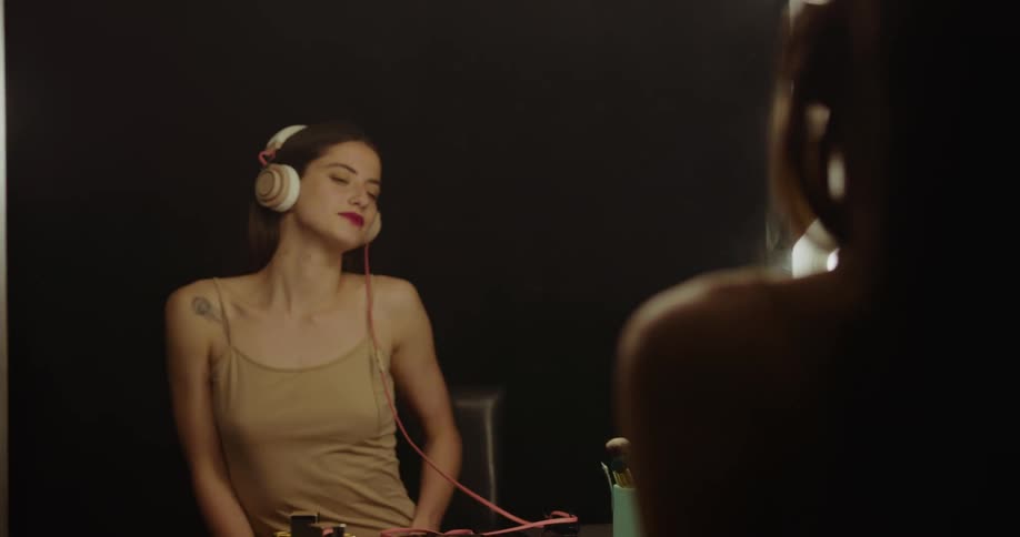 A woman sitting in at a vanity plugs her headphones into her smartphone before placing them on her head and dancing in the mirror
