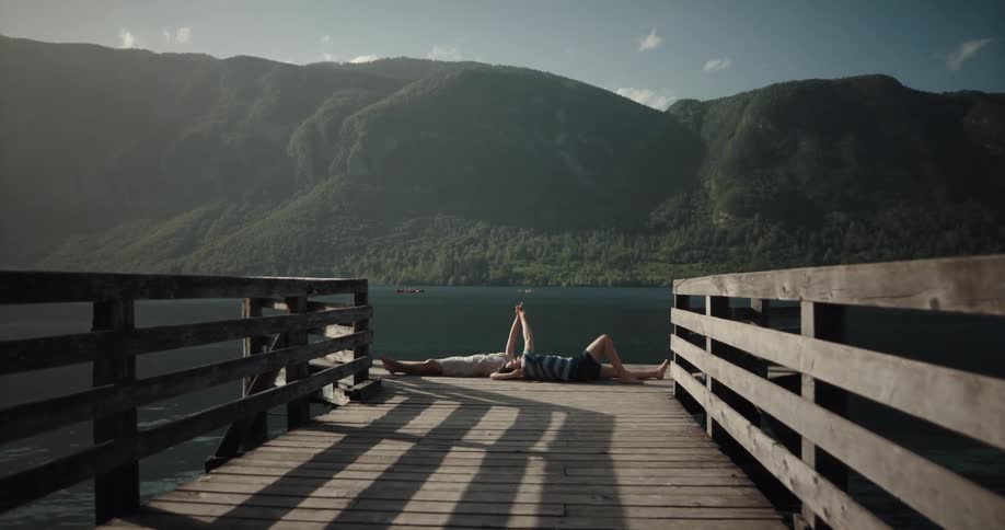 Two people lay on the end of a pier of a quiet lake