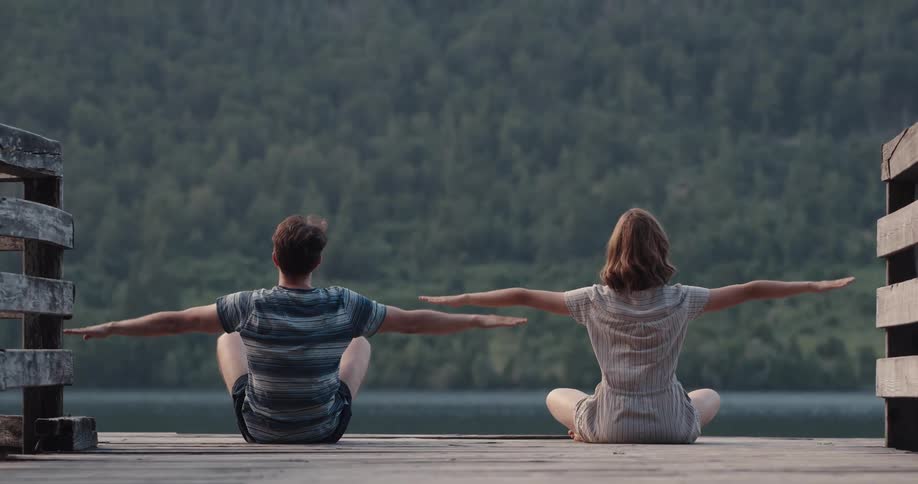 A man and woman sitting on a dock stretching with a river and trees in the background