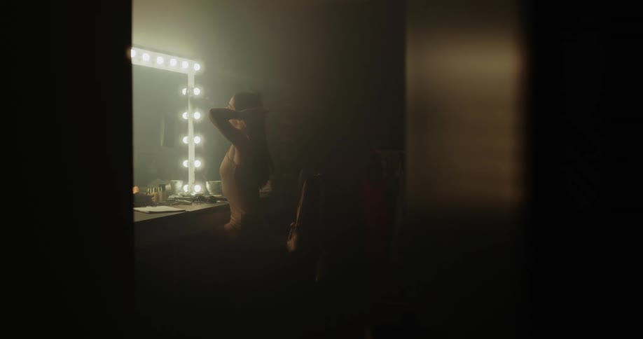 A woman brushes her long hair in a dressing room with lighted mirror