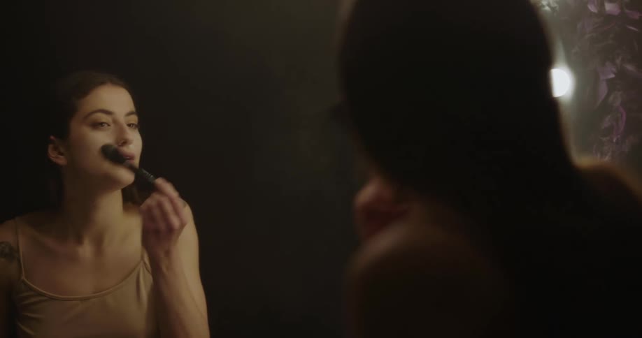 A young woman puts on powder makeup with a brush while looking in a large mirror