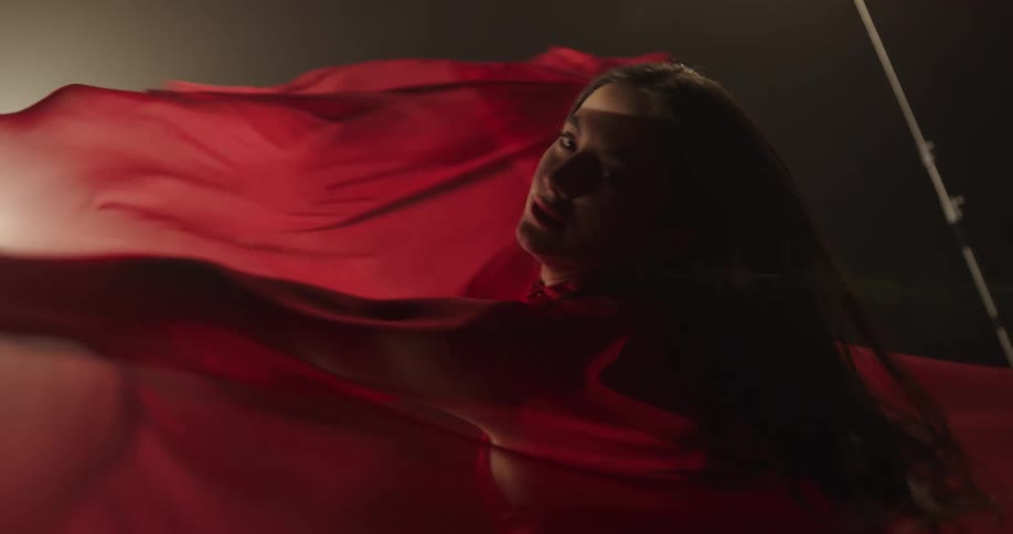 A woman waves a red cape over her head as she dances in a red dress under a spotlight
