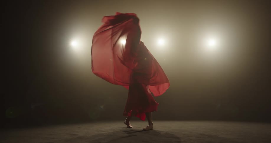 Woman wearing filmy red wrap dances barefoot alone in foggy room backed by row of lights