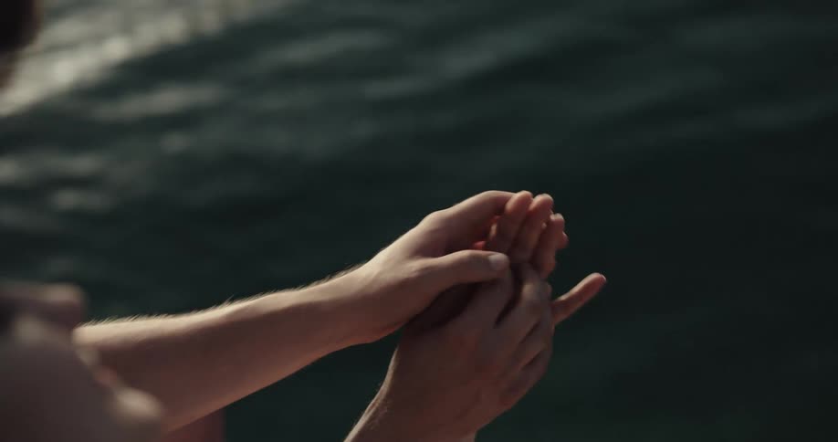 A man grabs his girlfriends hand and kisses it while they sit and dangle their feet over a lake