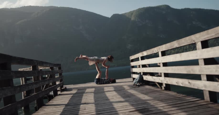 Man balances woman on his legs while lying on his back on pier across lake from mountain on misty day