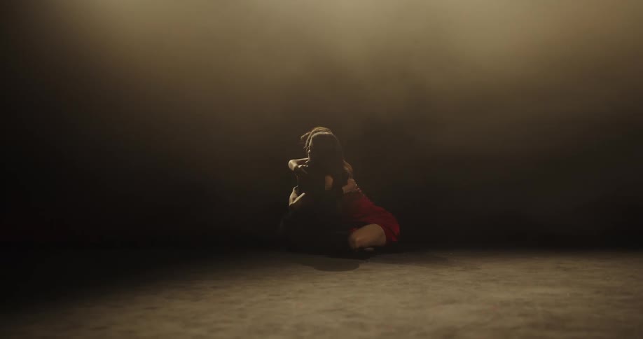 Two female dancers are seated on a poorly lit smoky stage hugging each other