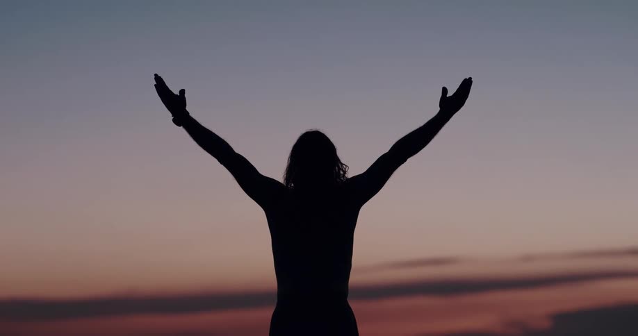 A person has their back turned away and is overlooking a beach and raising their arms in the air under a colorful sunset sky