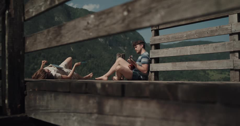 Two people sit and relax on the end of a pier with a book and a guitar