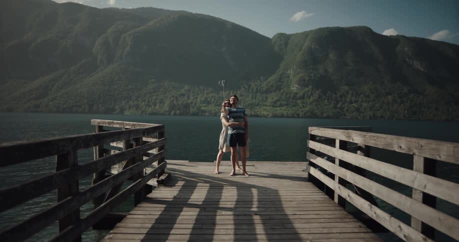 A man and woman are standing on a pier with their backs facing the water and taking a selfie
