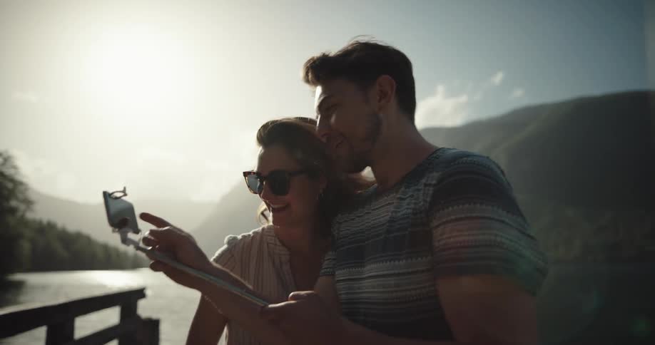 A man and a woman taking pictures with a smart phone and a selfish stick with a lake the sky and clouds in the background