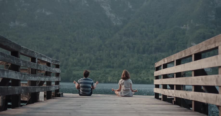 Girl and boy meditating on a wooden pier in a lake on the mountains