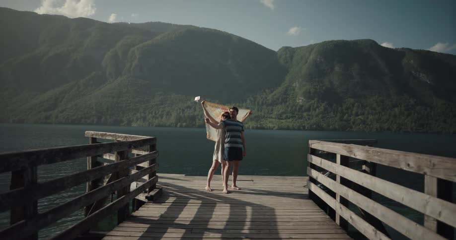 A man and woman are standing on a dock on a lake in the mountains while she holds up a scarf and they take selfies