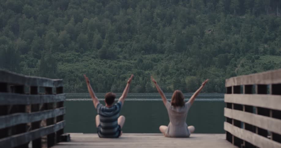 A man and woman stretch their arms while meditating on a pier
